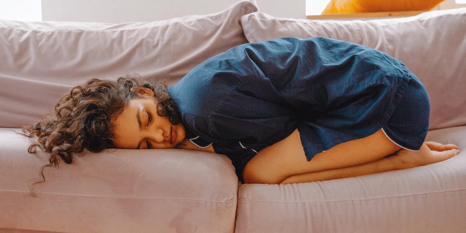 Woman is lying down on a sofa crouched over with her hands resting on her stomach in a foetus postion